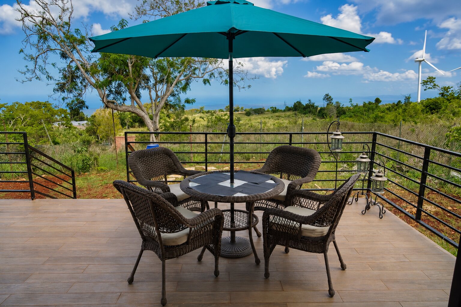 A patio deck with wicker seating under a teal umbrella, overlooking the hills and the sea